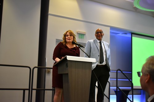 A woman with medium blonde hair in a red dress speaking at a podium, with a man in a blue suit jacket and black pants standing beside her and laughing.